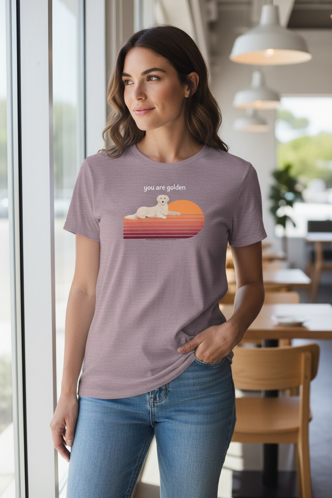 The Pupperfish™Woman wearing a t-shirt with a graphic design of a golden retriever and sunset, standing in a modern cafe.